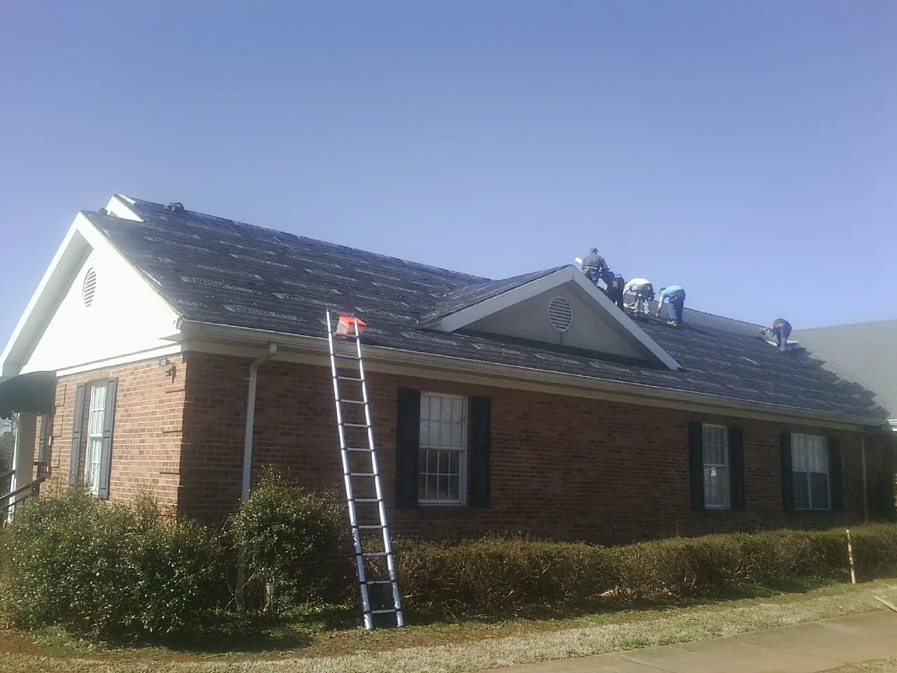 Crew tearing shingles off a brick ranch roof with a ladder set — Emergency Roof Repair work by our crew in Sanbornton