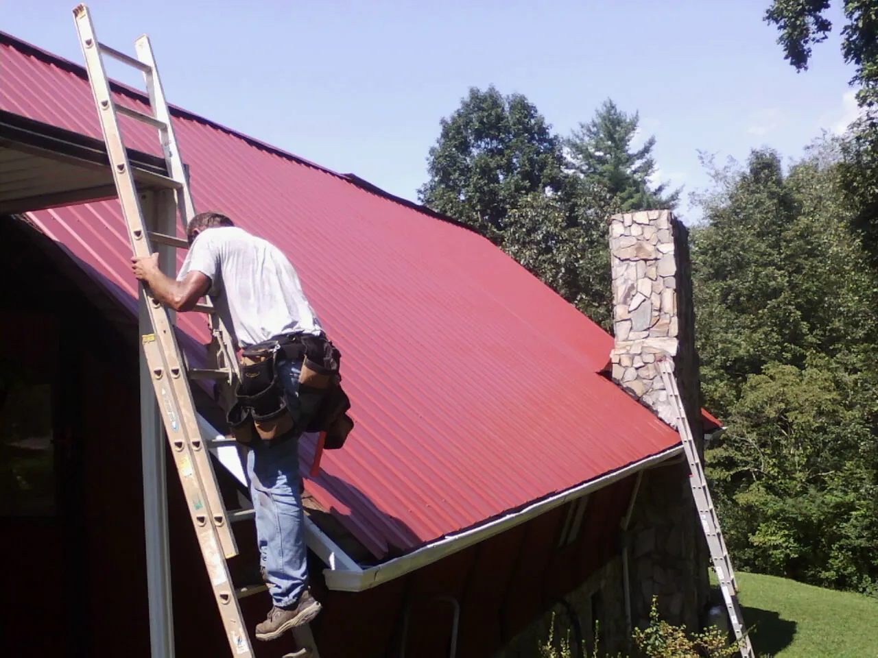 Roofer on a ladder beside a red metal roof and stone chimney — Soffit & Fascia Repair by Roofing Brothers in Sanbornton