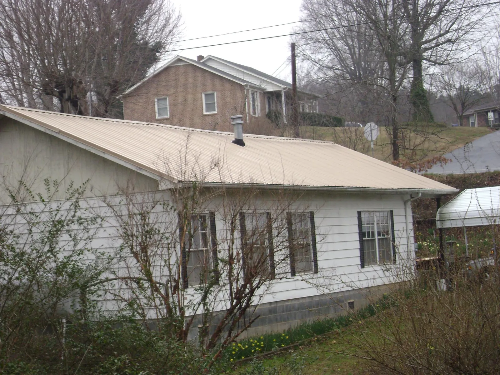 Tan metal roof on a white sided house set into a hillside — Commercial Roofing by Roofing Brothers in Sanbornton